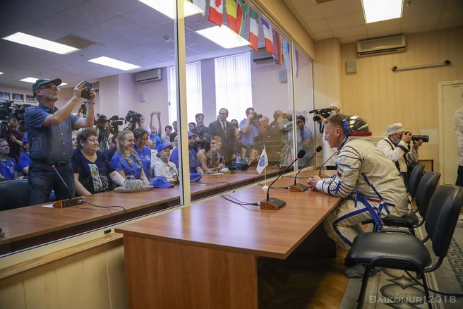 Уже одетые космонавты сидят за столом, который отделен от остальной части комнаты стеклом. До самого момента посадки в корабль экипаж изолируют от потенциально заразных окружающих
