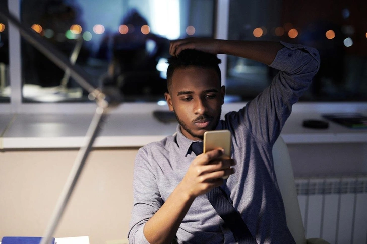 Офис-менеджер отдыхает в конце рабочего дня, сидя за столом и просматривая социальные сети на смартфоне | Источник: Dmitriy Shironosov / Alamy via Legion Media